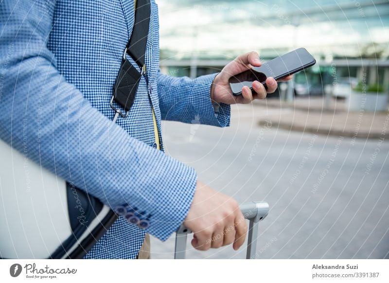 Stylischer Mann steht am Flughafen mit Smartphone und Koffer, blättert, textet, benutzt mobile Apps. Geschäftsreisende. Anwendung Ankunft Tasche Business