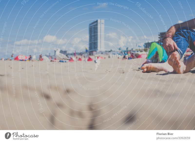 Barfuß bei schönem Wetter mit Hochgekrempelter Hose an einem belebten Strand sitzend Strandleben Gesellschaft Jeans Jeanshose Bekleidung blau lässig Textil Sand