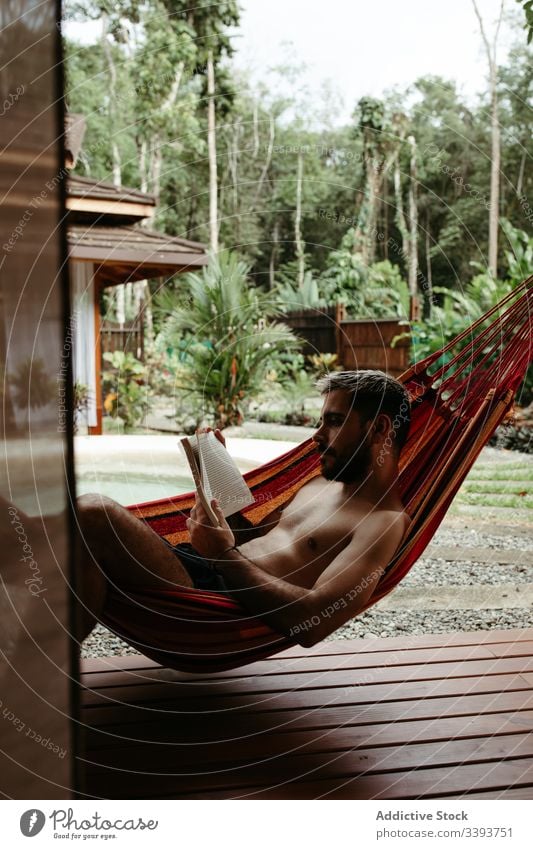 Junger männlicher Tourist in Bademode mit Buch auf Hängematte Mann Resort Hotel räkeln Kälte Beckenrand lesen exotisch tropisch Badeanzug sich[Akk] entspannen