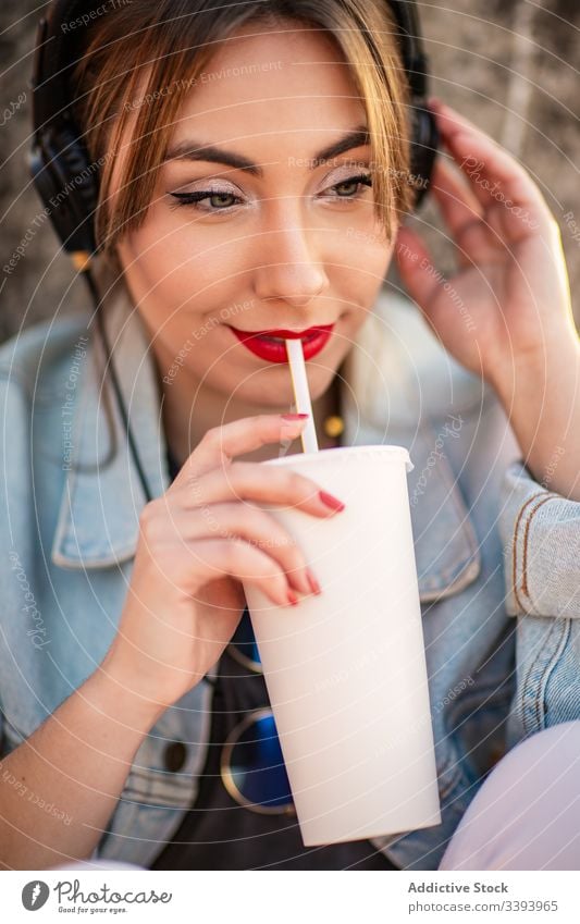 Wehmütige Frau benutzt Kopfhörer beim Entspannen ruhen sich[Akk] entspannen Natur sitzen Zaun lässig Getränk trinken benutzend Musik Stil Lifestyle Feiertag