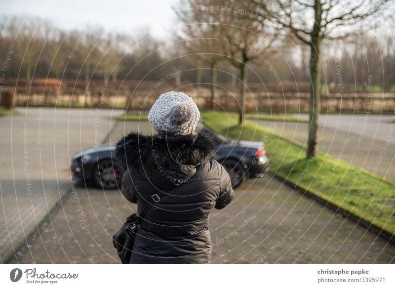 Frau in schwarzer Winterjacke und Mütze schaut auf schwarzen Roadster auf Parkplatz Jacke KFZ Tag parken roadster Sportwagen PKW Außenaufnahme Fahrzeug Cabrio