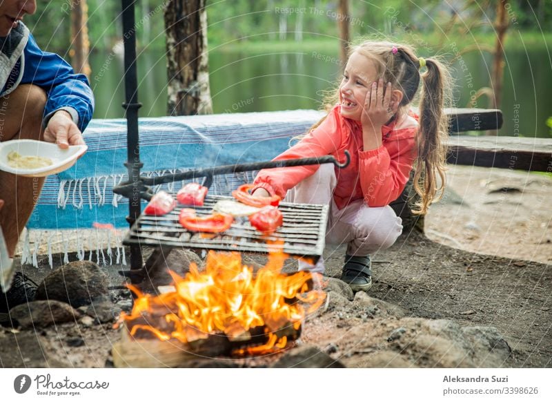 Mann und seine kleine Tochter grillen im Wald am felsigen Seeufer, machen ein Feuer, grillen Brot, Gemüse und Marshmallow. Familie erkundet Finnland. Skandinavische Sommerlandschaft.