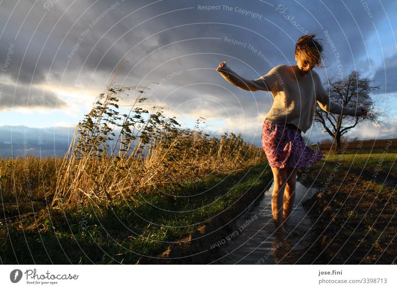 Frau steht in Pfütze im Feld Junge Frau Rock feminin Tag Wiese Acker Wolken Gewitterwolken windig Natur Landschaft Barfuß düster Sonnenlicht Ganzkörperaufnahme