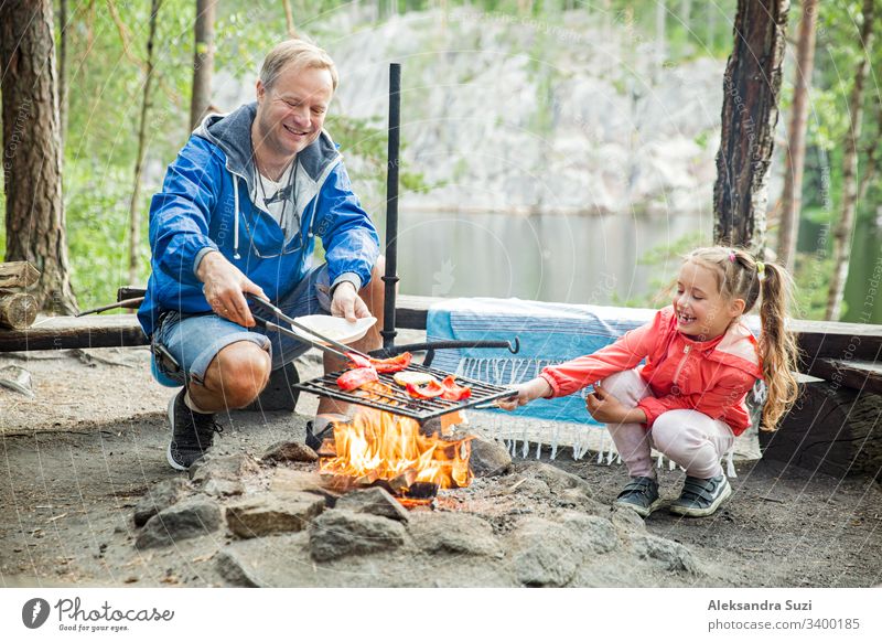 Mann und seine kleine Tochter grillen im Wald am felsigen Seeufer, machen ein Feuer, grillen Brot, Gemüse und Marshmallow. Familie erkundet Finnland. Skandinavische Sommerlandschaft.