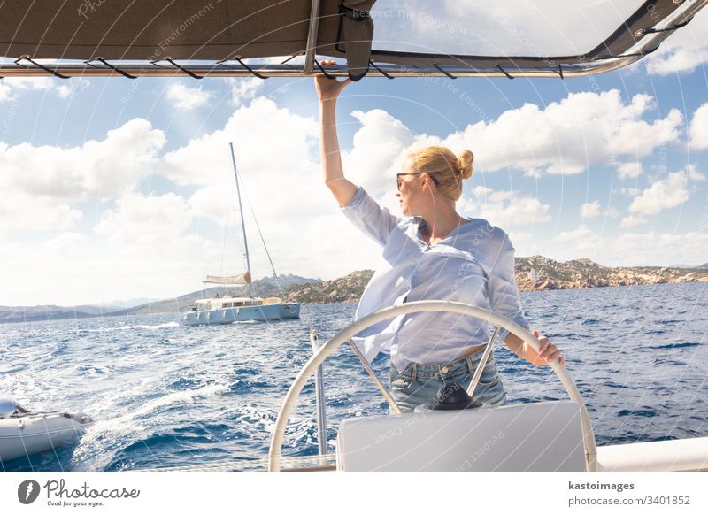 Attraktive blonde Skipperin, die das schicke Katamaran-Segelboot an einem sonnigen Sommertag auf ruhigem blauen Meerwasser steuert. nautisch Lenkrad Boot Frau