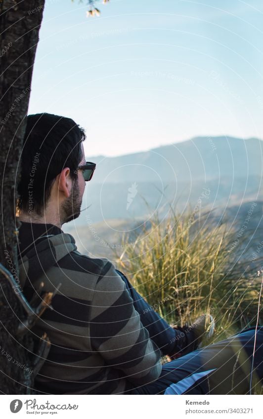 Seitenansicht eines Mannes mit Sonnenbrille, der an einem sonnigen Tag auf dem Gras in der Nähe eines Baumstammes sitzt, mit den Bergen im Hintergrund. sitzen