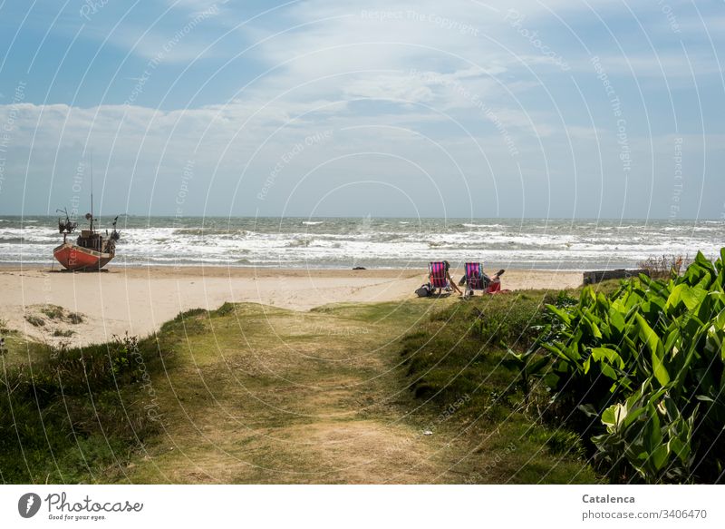 Zwei Personen bei windigem Wetter am Strand sehen der Brandung zu, links von ihnen liegt ein Fischerboot im Sand Strandleben Wasser Spaß Farbfoto Himmel Sommer