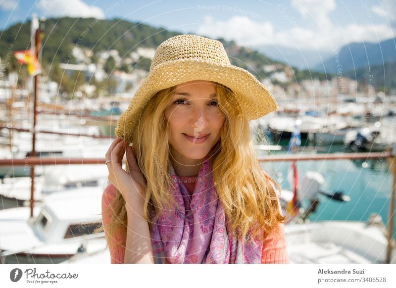 Porträt einer jungen Frau mit hellem lockigem Haar und Strohhut, die lächelnd Sonne und Wind genießt. Sonniger Hafen mit Booten und Jachten, grüne Berge im Hintergrund. Lebensfreude, fröhliche Reisende,