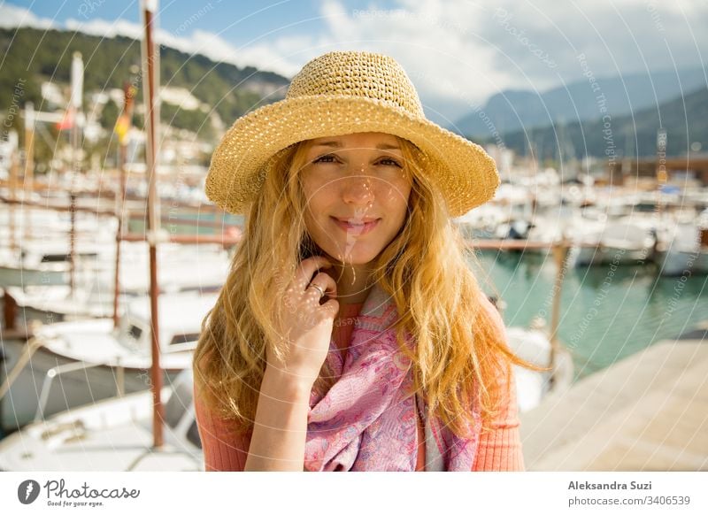 Porträt einer jungen Frau mit hellem lockigem Haar und Strohhut, die lächelnd Sonne und Wind genießt. Sonniger Hafen mit Booten und Jachten, grüne Berge im Hintergrund. Lebensfreude, fröhliche Reisende,