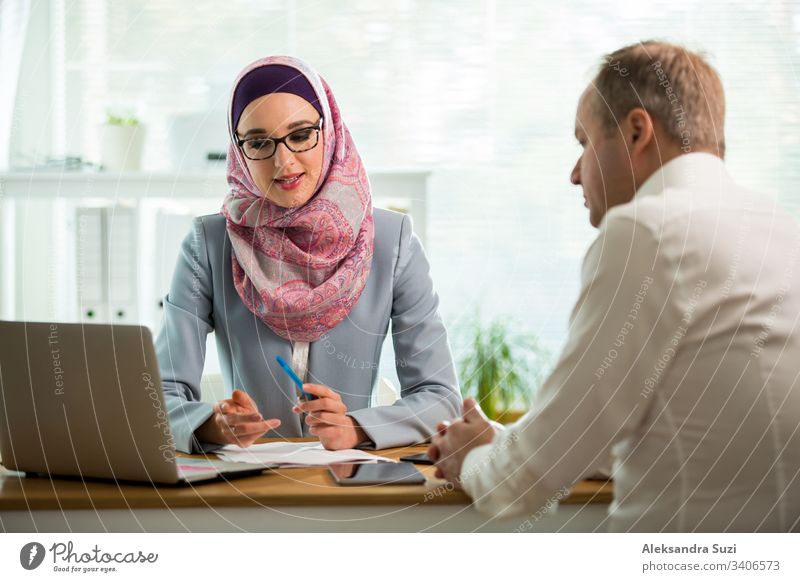 Treffen der Mitarbeiter im Büro. Stilvolle Frau im Hijab, die sich am Schreibtisch mit einem Mann in einem modernen weißen Büro unterhält. Muslimische Geschäftsfrau mit Brille interviewt Mann.