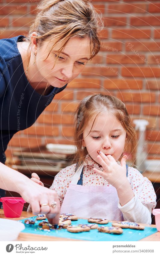 Kleines Mädchen mit Hilfe ihrer Mutter, das gebackene Kekse mit bunten Streuseln und Puderzucker verziert. Kind nimmt am Backworkshop teil. Backkurse für Kinder, aufstrebende kleine Köche. Kochen lernen. Kombinieren und Rühren von vorbereiteten Zutaten