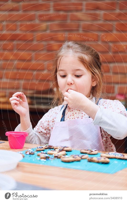 Kind nimmt an Backworkshop teilKleines Mädchen probiert selbst gebackene Kekse. Sie verziert ihre gebackenen Kekse mit bunten Streuseln und Puderzucker. Kind, das am Backworkshop teilnimmt. Backkurse für Kinder, aufstrebende kleine Köche. Kochen lernen