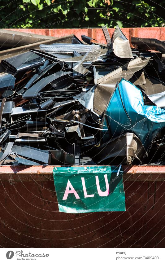 Ein Schrottcontainer voller, teils buntem, Aluschrott eines Dachdeckers. Vorne eine Tafel mit der Beschriftung Alu Aluminium Sammelsurium kunterbunt unbrauchbar