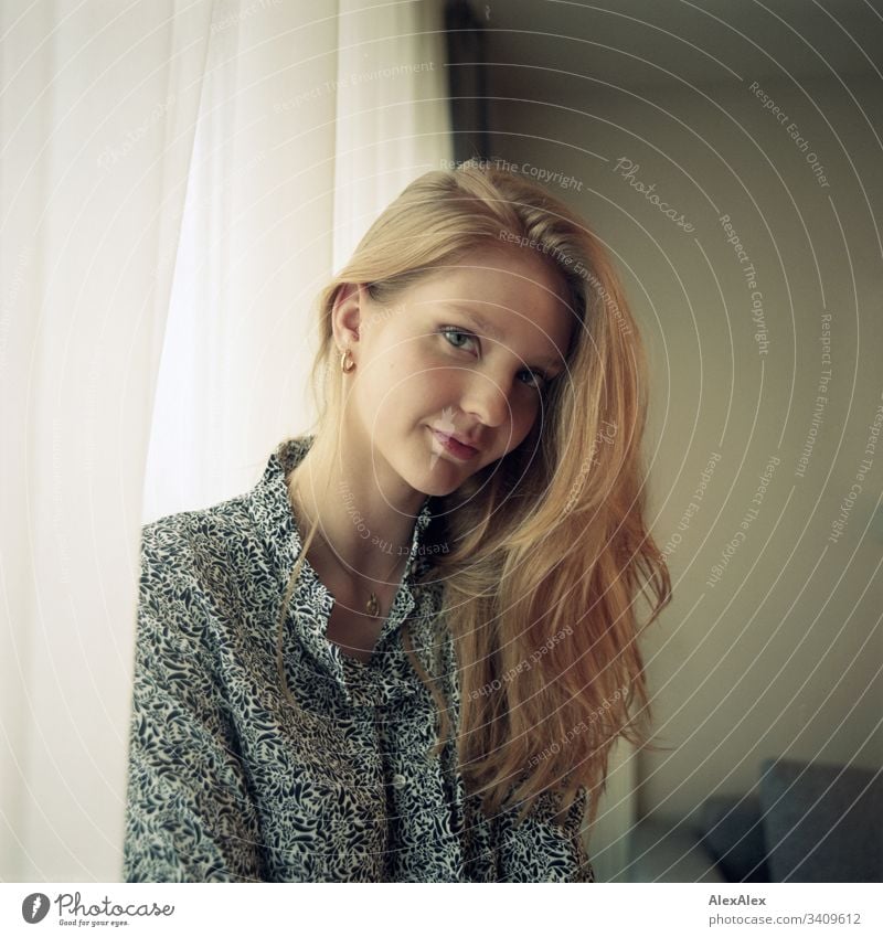 Portrait einer jungen Frau neben dem Fenster an der Gardine Mädchen blond schön schlank anmutig elegant lifestyle Wohnen Wohnung zu Hause Bluse Muster