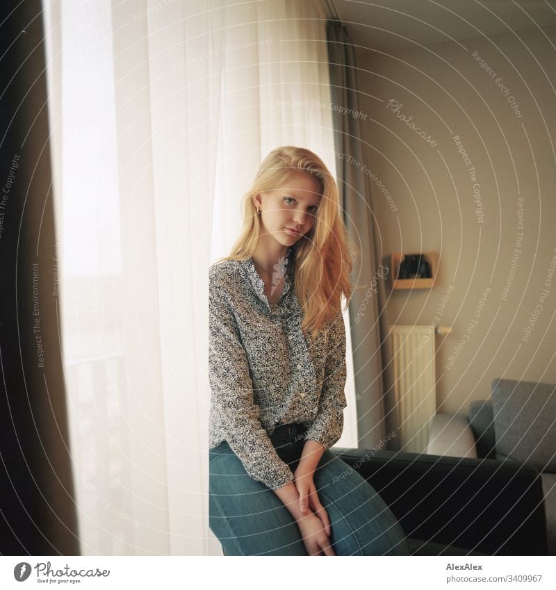 Portrait einer jungen Frau neben dem Fenster an der Gardine Mädchen blond schön schlank anmutig elegant lifestyle Wohnen Wohnung zu Hause Bluse Muster