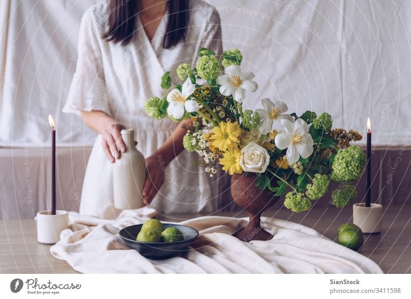 Frau hält Flasche. Stilleben mit Blumenkonzept Tisch jung Kaukasier Halt Beteiligung Hände Hand Kleid weiß Vase brünett Kerzen Kalk Blumenstrauß