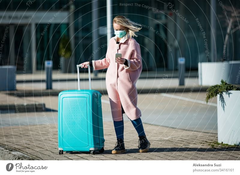 Frau mit Schutzmaske steht am Flughafen-Eingang mit Smartphone und großem Gepäckkoffer, blättert, simst, benutzt mobile Apps. Sicheres Reisen. Ankunft plaudernd