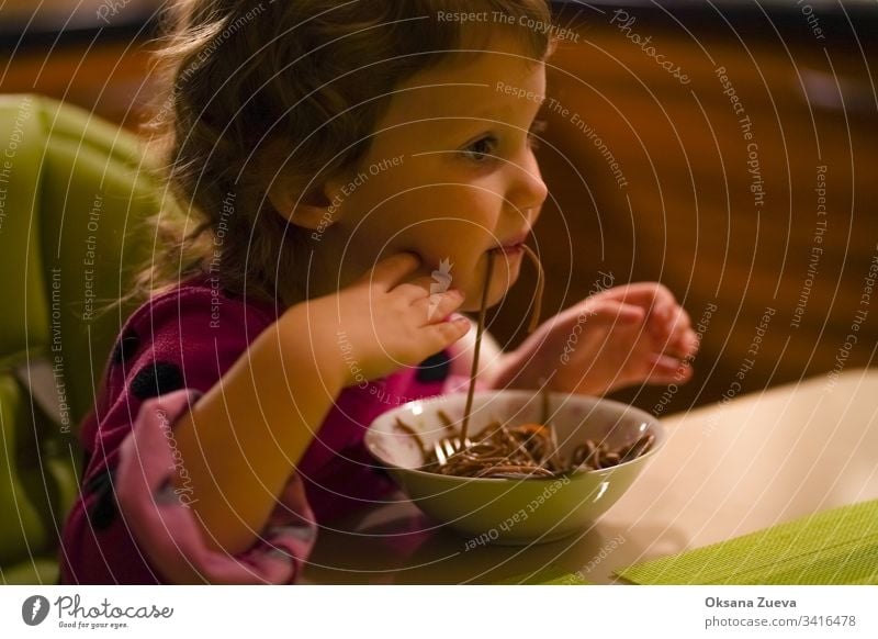 Das gelockte kleine Mädchen mit Appetit isst Buchweizennudeln, Spaghetti. Das Konzept des guten Appetits, leckeres selbstgemachtes Essen. bezaubernd