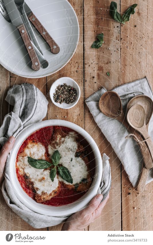 Nicht erkennbare Person, die eine Schüssel voll Huhn mit Käse und Basilikum in Tomatensauce auf den Tisch stellt Speise Utensil Braten Hähnchen Saucen