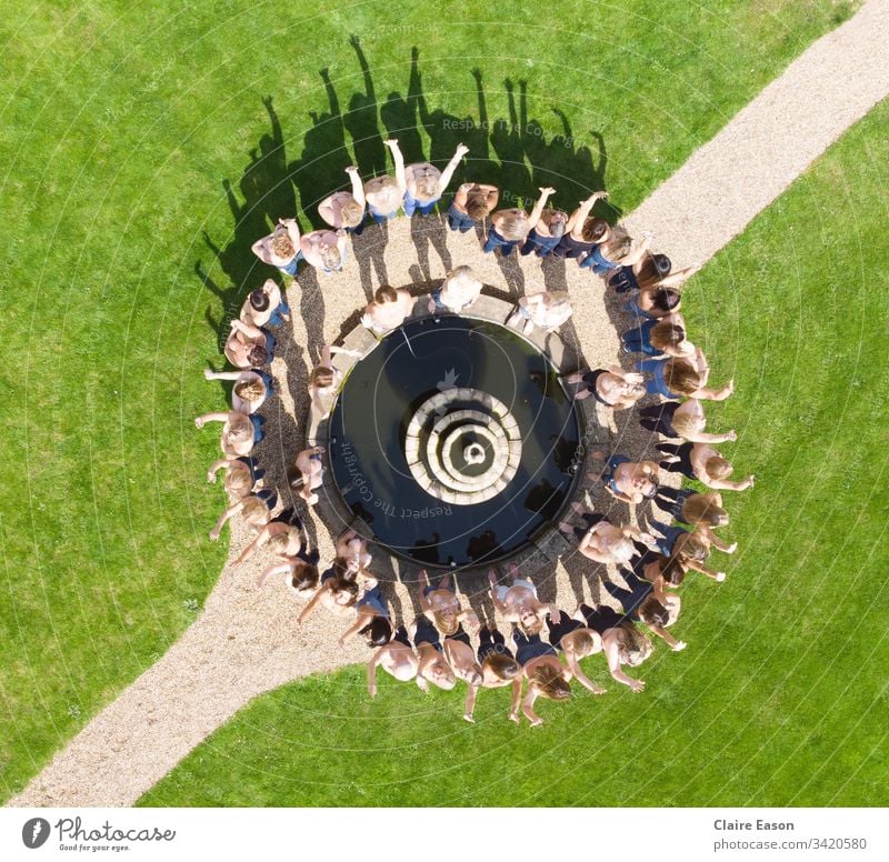 Luftaufnahme von etwa 50 Frauen in Jeans, um einen Zierbrunnen herum, mit einem diagonalen Weg und grüner Umgebung. Brustbewusstsein Schwestern Brustkrebs