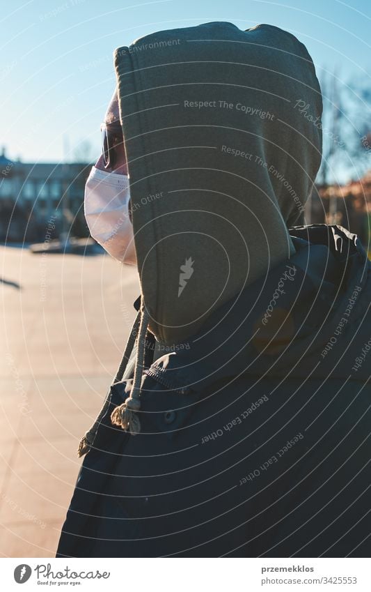 Junger Mann mit Kapuze, der die Gesichtsmaske trägt, um eine Virusinfektion zu vermeiden und die Ausbreitung der Krankheit zu verhindern. Spaziergang im Stadtzentrum. Schutz vor einer Virusinfektion. Echte Menschen, authentische Situationen