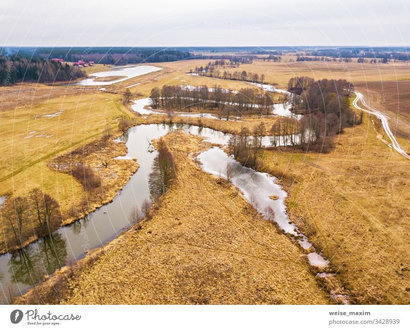 Kleiner Fluss mit ausgetrockneten Bäumen ohne Blätter im Winter / Anfang Frühling - Luftaufnahme. Launisches, bedecktes Wetter Bach strömen Straße Wegbiegung