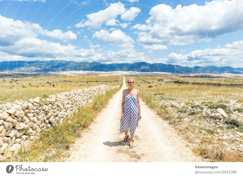 Kaukasische junge Frau in Sommerkleidung hält einen Strauß Lavendelblüten in der Hand, während sie im Sommer durch die trockene, felsige Landschaft der kroatischen Mittelmeerküste auf der Insel Pag spaziert