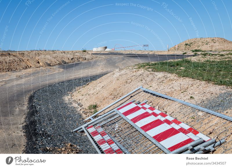 Blick in ein Baugebiet Neubaugebiet Baustelle ruhend rotweiß Absperrung Absperrgitter kurvenreich Straße brachliegend Blauer Himmel