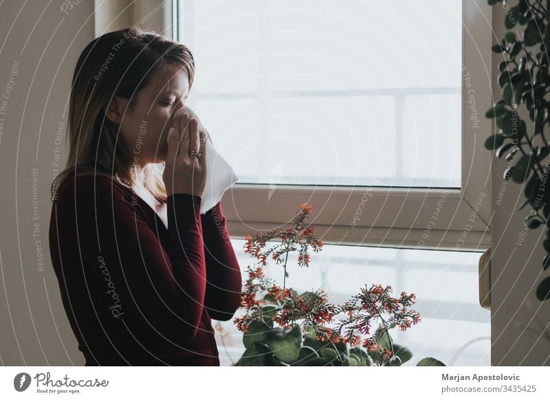 Junge kranke Frau schnäuzt sich am Fenster in ein Taschentuch 30s Erwachsener Allergie allein Appartement blasend Pflege lässig Kaukasier kalt Ansteckung dunkel