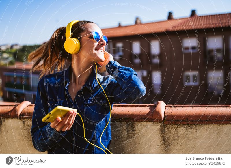 junge Frau, die über ein Mobiltelefon und ein gelbes Headset Musik hört. Spaß und Lebensstil hören Handy Technik & Technologie Terrasse im Freien sonnig