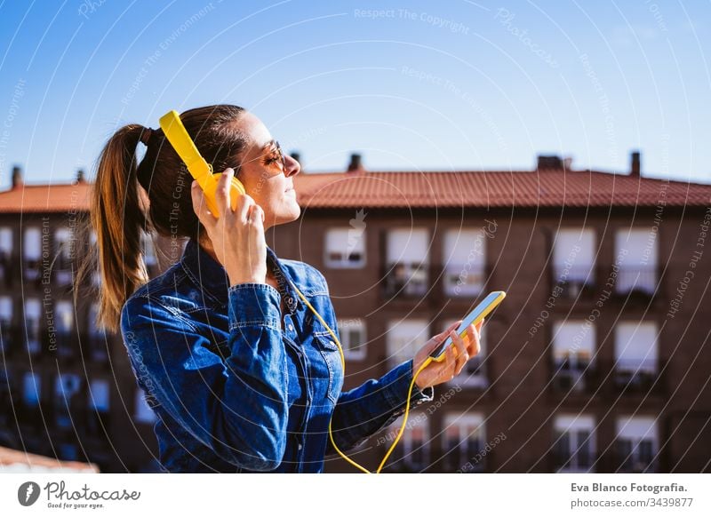 junge Frau, die über ein Mobiltelefon und ein gelbes Headset Musik hört. Spaß und Lebensstil hören Handy Technik & Technologie Terrasse im Freien sonnig