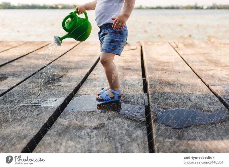 Ein kleines Mädchen spielt mit der Gießkanne einer Holzbrücke. Frühling und Sommer. Gartenarbeit. grüne Gießkanne Bewässerung Dose Wasser Baby wenig Kind