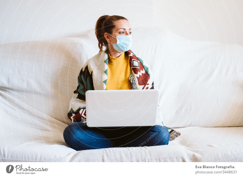 junge Frau, die zu Hause am Laptop arbeitet, auf der Couch sitzt und eine Schutzmaske trägt. Das Konzept "zu Hause bleiben" während des Coronavirus covid-2019