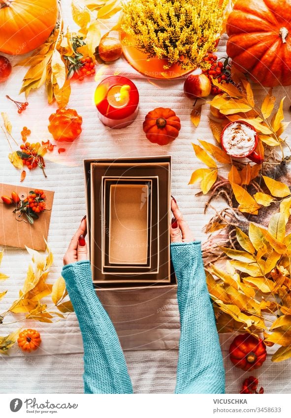 Frauenhände in blauem Pullover halten leere umweltfreundliche Bastelkartons mit verschiedenen Kürbissen, Herbstblättern, Kerzen und Cappuccino in rotem Becher auf weißem Deckenhintergrund auf dem Schreibtisch. Ansicht von oben. Flach gelegt