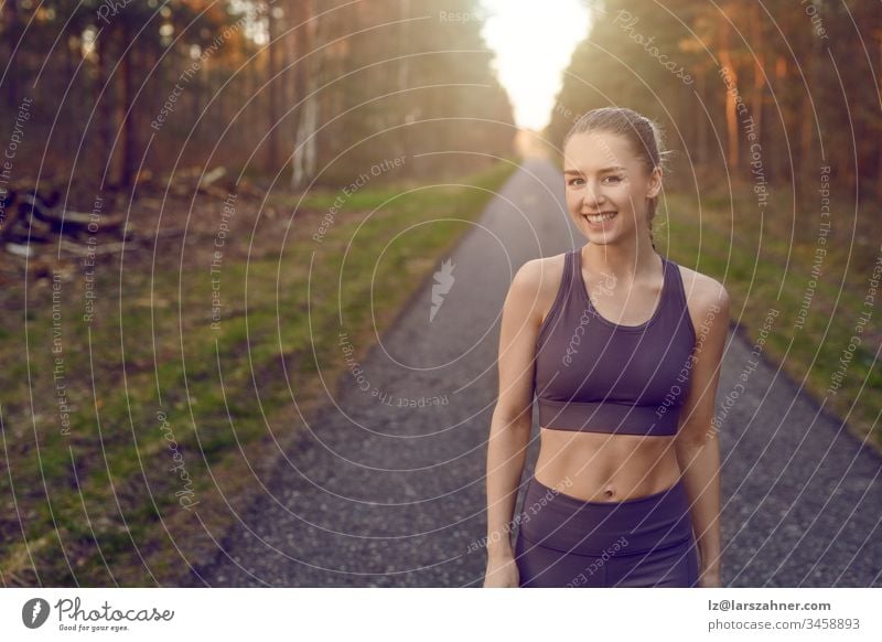 Lächelnde, sportlich-sportliche junge Frau, die auf einer geteerten Bahn durch Wälder, die vom warmen Schein der Sonne beleuchtet werden, in einem gesunden, aktiven Lebensstil trainiert