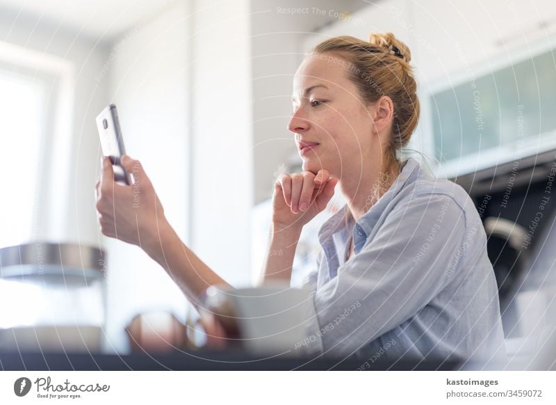 Junge, lächelnde, fröhliche, zufriedene Frau, die zu Hause in der Küche sitzt und soziale Medien auf dem Handy nutzt, um mit ihren Lieben zu plaudern und zu stylen. Bleiben Sie zu Hause, sozialer distanzierender Lebensstil.