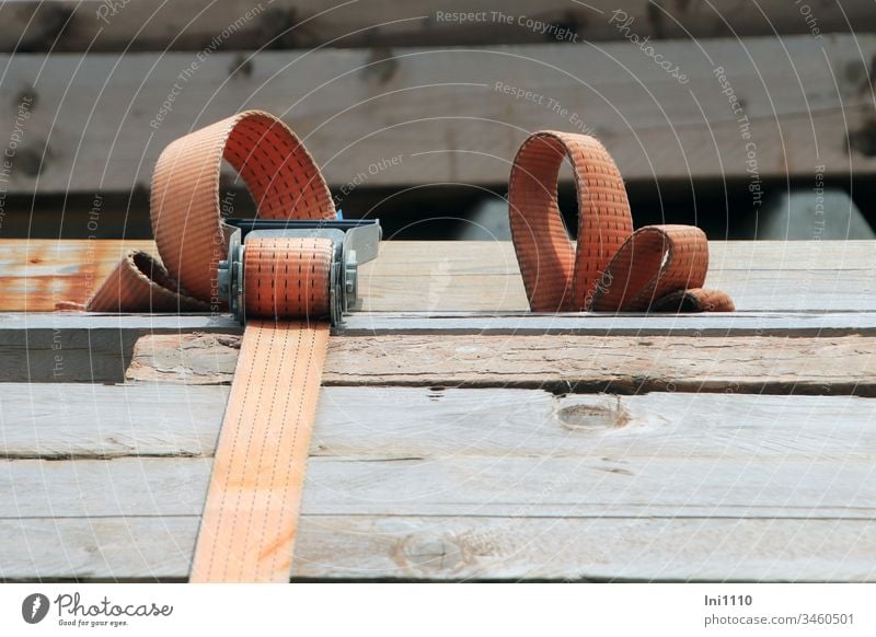 Spanngurt in orange mit Ratsche umschließt Stapel Holzbohlen Gurtende rollt sich zu kleinen Schleifen Sicherheit starker Halt Schnalle Ösen Befestigung