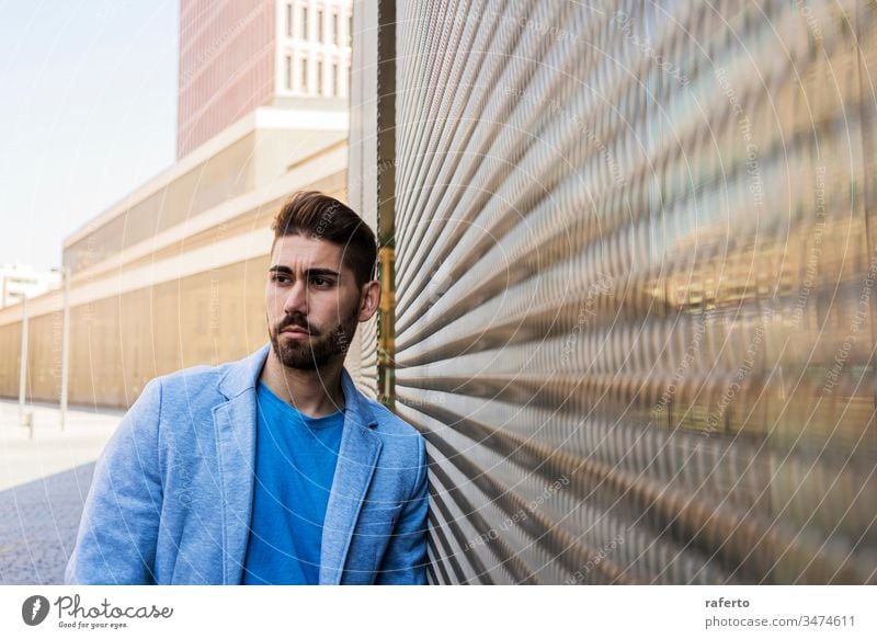 Junger bärtiger Mann, Modell der Mode, im städtischen Hintergrund, in legerer Kleidung, während er an einer Wand lehnt und zur Seite schaut cool Stil Porträt