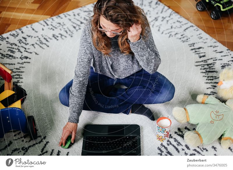 Frau sitzt im Schneidersitz im Kinderzimmer mit Spielzeug und arbeitet am Laptop Homeoffice sitzen arbeiten stayhome Teppich Computer Arbeit Erwachsener Büro