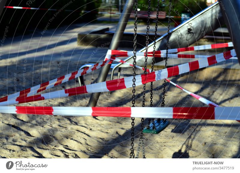 Corona Thoughts | Spielen verboten! Rot weißes Absperrband kreuz und quer durch eine Kinderschaukel auf einem Kinderspielplatz mit Sonnenlicht und Schatten