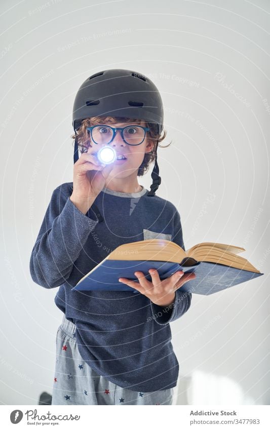 blondes Kind im Schlafanzug mit einem Helm, einer Taschenlampe und einem Buch, das Forschung spielt Kinder Schutzhelm Spiel behüten Fröhlichkeit Genuss spielen