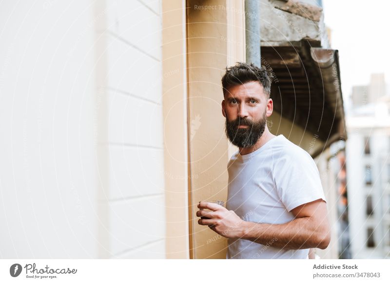 Bärtiger Typ mit heißem Getränk auf dem Balkon ruhend Mann trinken Morgen lässig stehen modern heimwärts Appartement männlich Vollbart Erwachsener gutaussehend