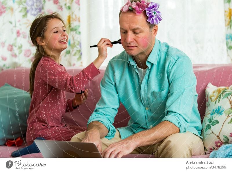 Kind spielt und stört Vater, der fern von zu Hause arbeitet. Kleines Mädchen, das mit einem Pinsel Schminke aufträgt. Mann sitzt mit Laptop auf der Couch. Familie verbringt Zeit miteinander im Haus.