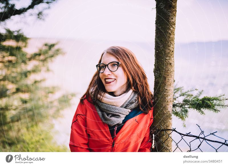 Junge Frau mit roter Jacke lehnt entspannt an einem Baum und lächelt mit der Sonne um die Wette spazierengehen Spaziergang Abenteuer Gesundheit Abstand Hügel