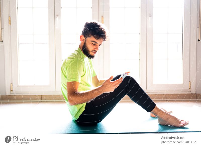 Nachdenklicher, entspannter Sportler, der ein Smartphone benutzt, während er sich nach dem Training auf einer Sportmatte im Leichtathletikraum ausruht Unterlage