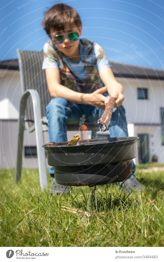 Schulkind sitzt im Garten bei einer kleinen Feuerstelle grillen Griller Grillsaison Grillplatz Grillen Außenaufnahme Kind Junge halten Ast brennen anzünden