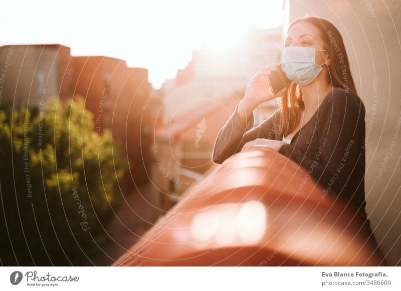 junge Frau zu Hause auf einer Terrasse, die eine Schutzmaske trägt, ein Mobiltelefon benutzt und einen sonnigen Tag genießt. Coronavirus Covid-19-Konzept Handy