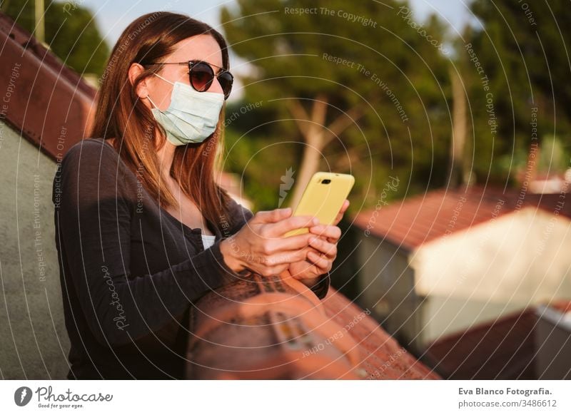 junge Frau zu Hause auf einer Terrasse, die eine Schutzmaske trägt, ein Mobiltelefon benutzt und einen sonnigen Tag genießt. Coronavirus Covid-19-Konzept Handy