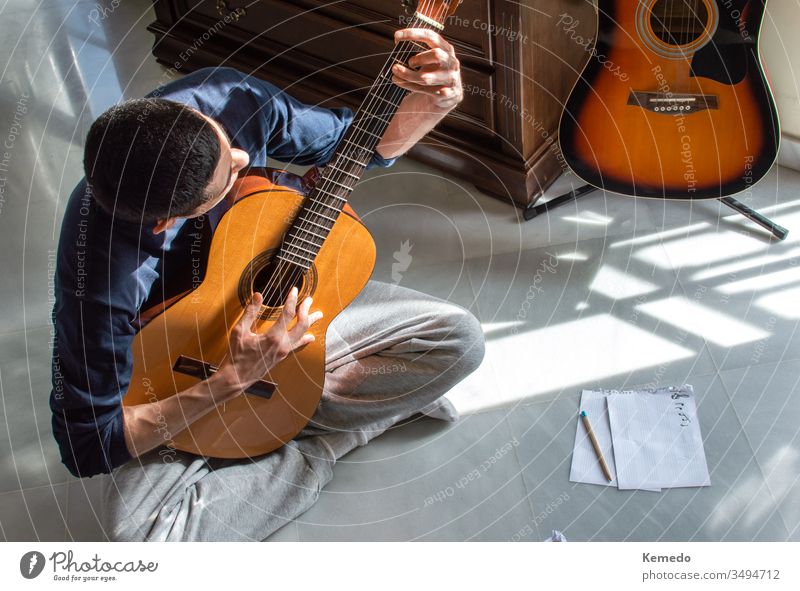 Mann, der an einem sonnigen Tag zu Hause in der Nähe eines hellen Fensters Gitarre spielt und Musik komponiert. Gelegenheitsmusiker, der auf dem Boden sitzt und Gitarre spielt.