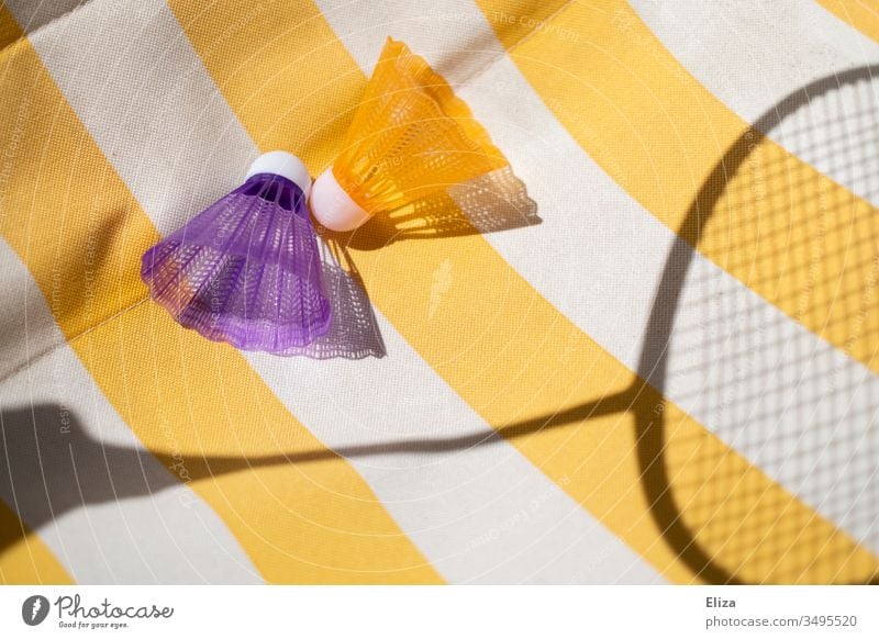 Zwei bunte Federbälle mit dem Schatten eines Federballschlägers auf einem gelb weiß gestreiftem Hintergrund sommerlich Sport Hobby spielen Badminton